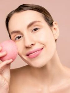 Close-up of a woman smiling while using a pink facial cleansing sponge for skincare.